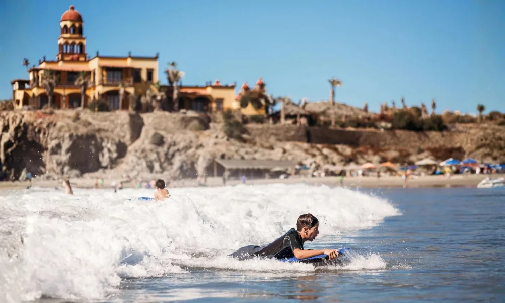Surfer riding a wave at Cerritos Beach, with the rocky point and a hilltop building behind