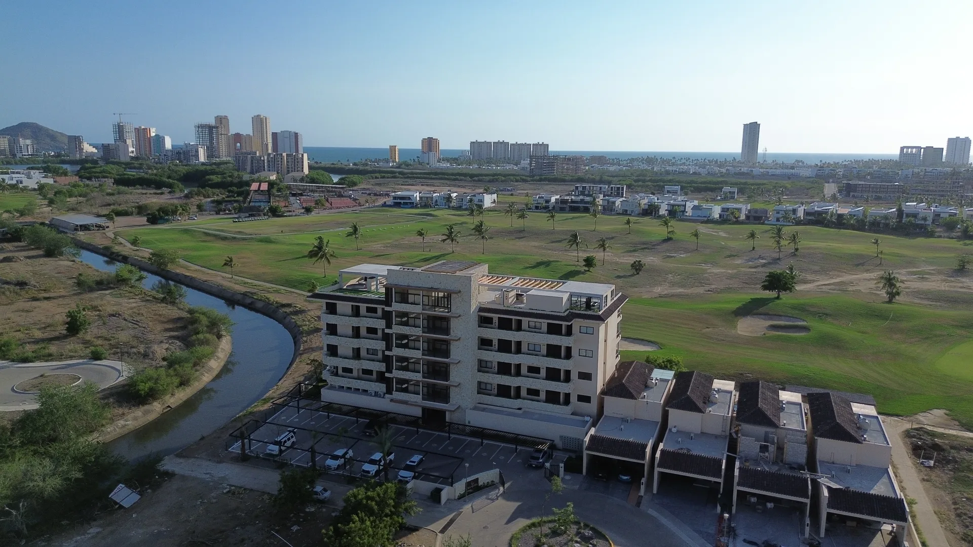 Vista aérea de Mangata Golf & Living en Marina Mazatlán: la torre de departamentos y las villas junto al campo de golf, con la ciudad y el mar al fondo